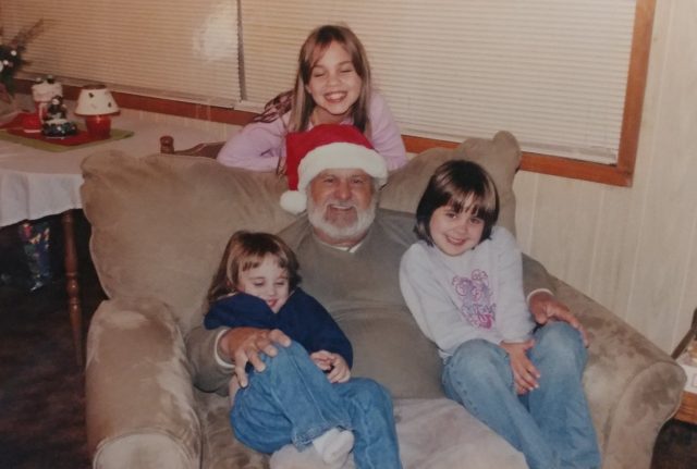 A senior man wearing a Santa hat sits with a grandchild in each arm. A third, older grandchild stands behind the armchair, and all smile warmly.
