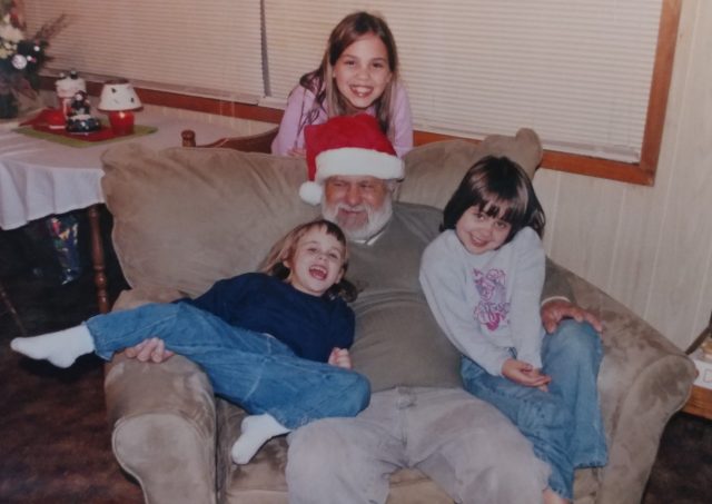 A senior man wearing a Santa hat sits with a grandchild in each arm. A third, older grandchild stands behind the armchair, and all smile warmly. 