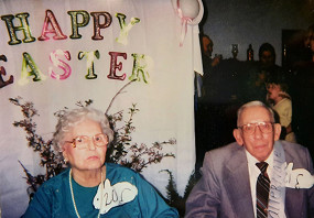 Elderly man and woman smiling in front of a Happy Easter photo backdrop.