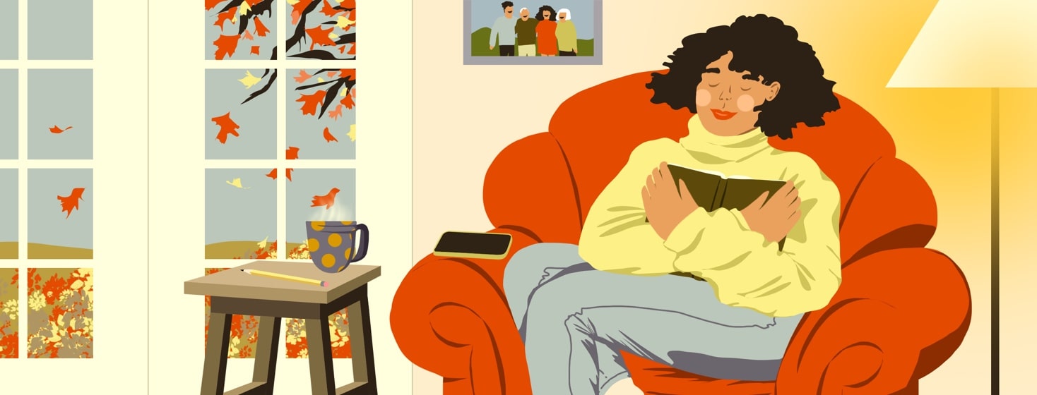 A woman sits in a comfy chair near a window where autumn leaves are falling. She has an open journal pressed to her chest and is smiling with her eyes closed.