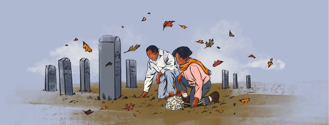 A man and woman crouch on the ground, laying flowers at a tombstone in a cemetery. They are dressed for fall and colored leaves blow past them.
