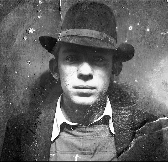 A black and white photo, worn and damaged with age but still striking, showing a young adult man in a stylish outfit and fedora.