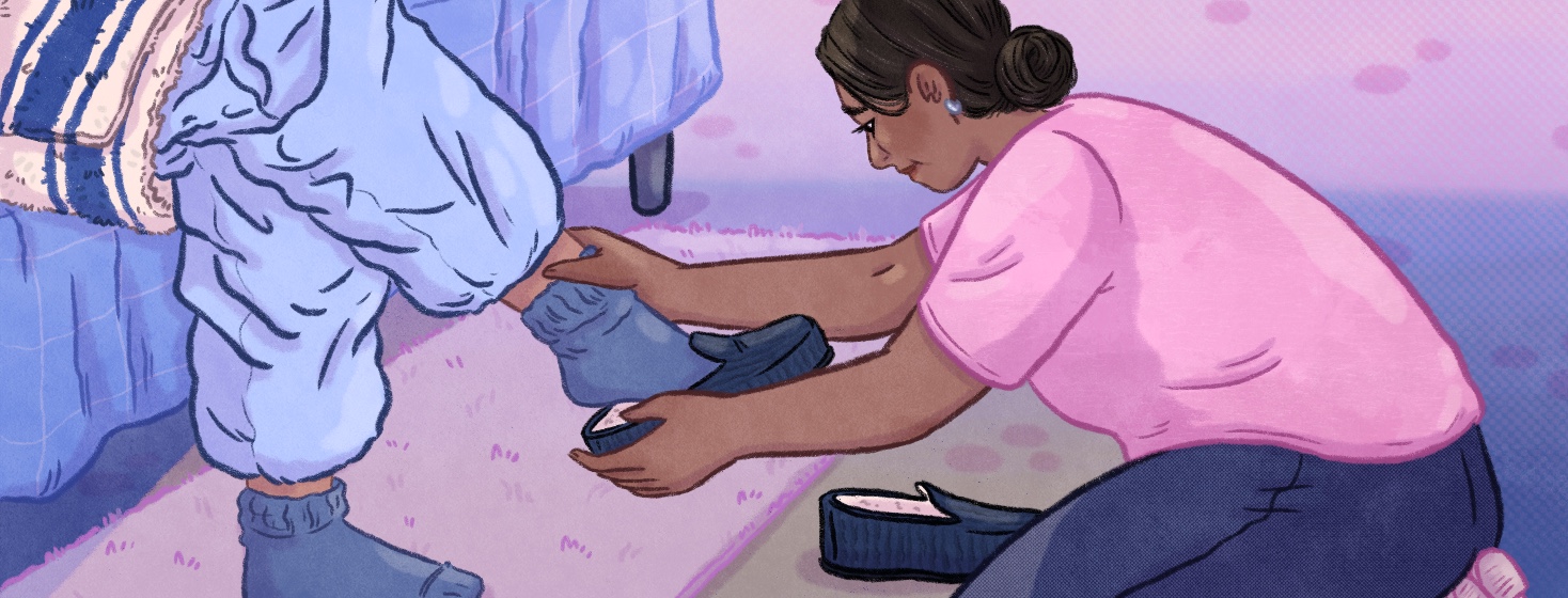 A woman gently puts one shoe on a person wearing loose sweatpants and a towel sitting on the edge of a bed with the second shoe next to her on the floor