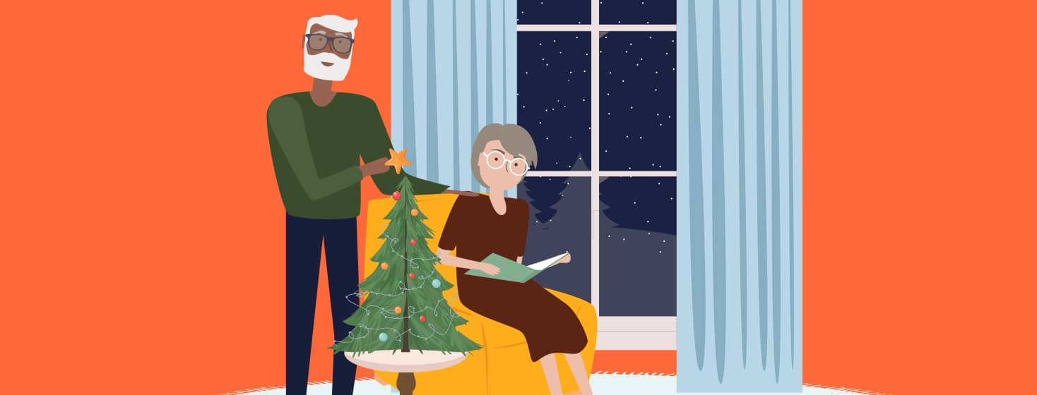 A senior man in glasses helps decorate a tabletop Christmas tree while a senior woman sits in a recliner looking at a book.