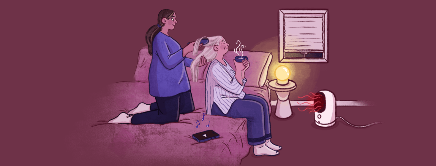 A woman kneels on a bed brushing the hair of a senior woman who drinks a warm drink; a phone on the bed plays music and there is a space heater on the floor