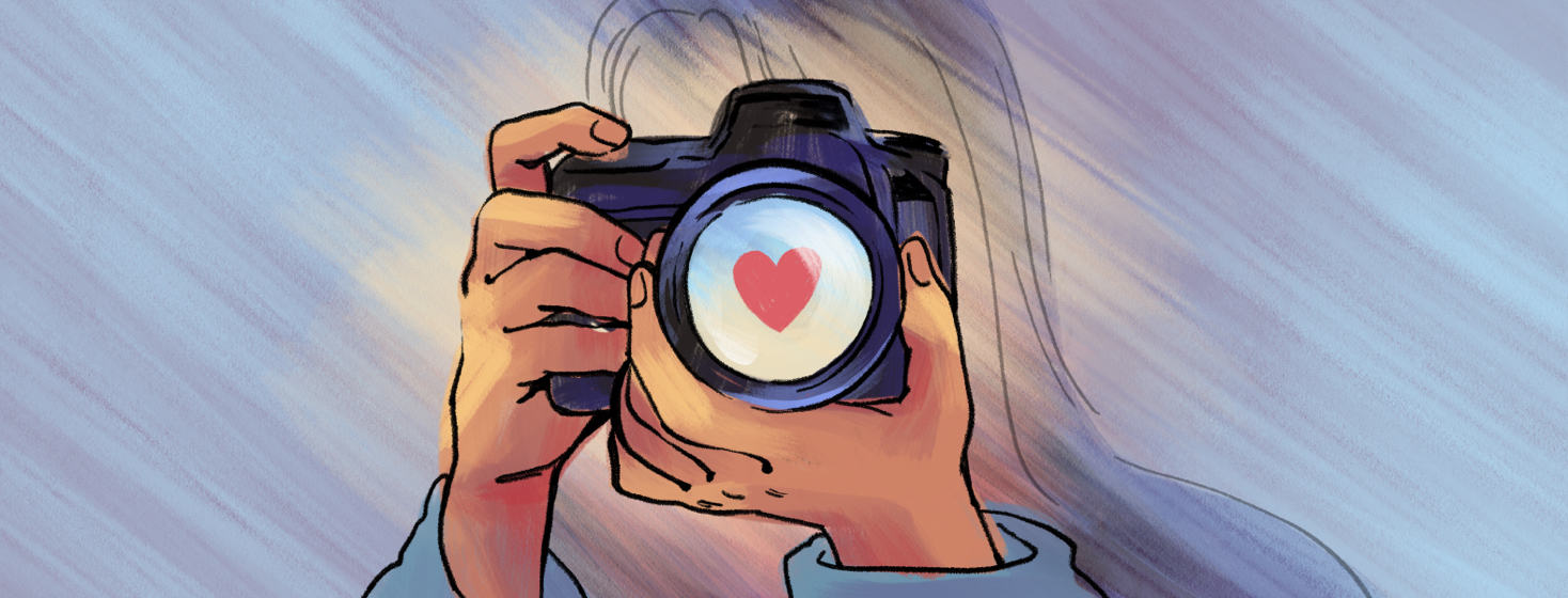 A person holds a camera with a heart shape reflected in the lens.